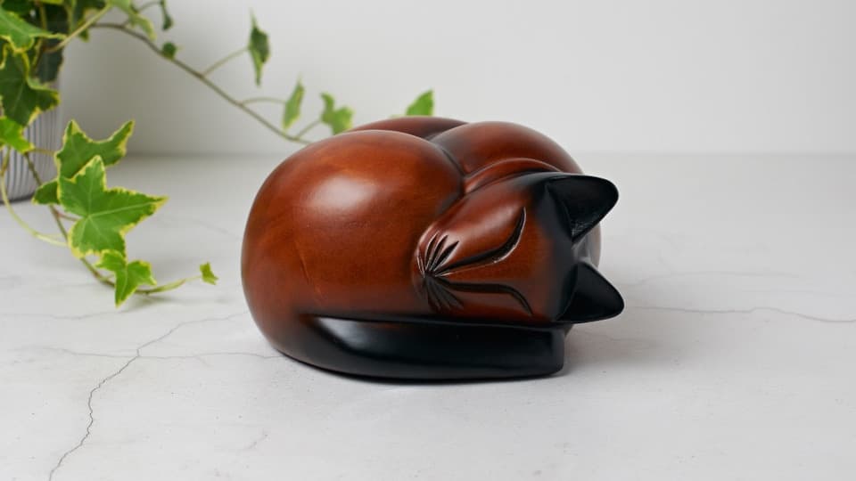 A wooden urn of a sleeping cat, curled round with its head snuggled into its tail. The urn is stained a red-brown on its body and face, and black on its ears and tail